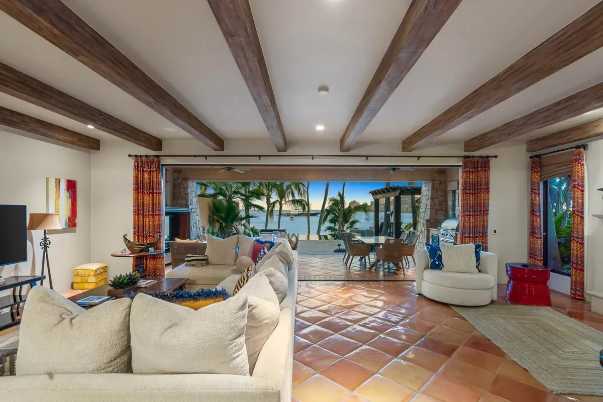 Living room with beige sectional sofa, white armchair, TV, colorful curtains, and view of a patio with palm trees and water outside.