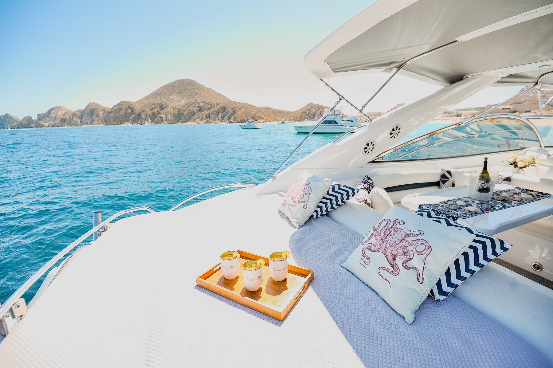 A luxurious boat deck with pillows featuring octopus designs, a tray with four cups of a beverage, and a table with flowers and wine, overlooking the ocean with boats and rocky islands in the distance.