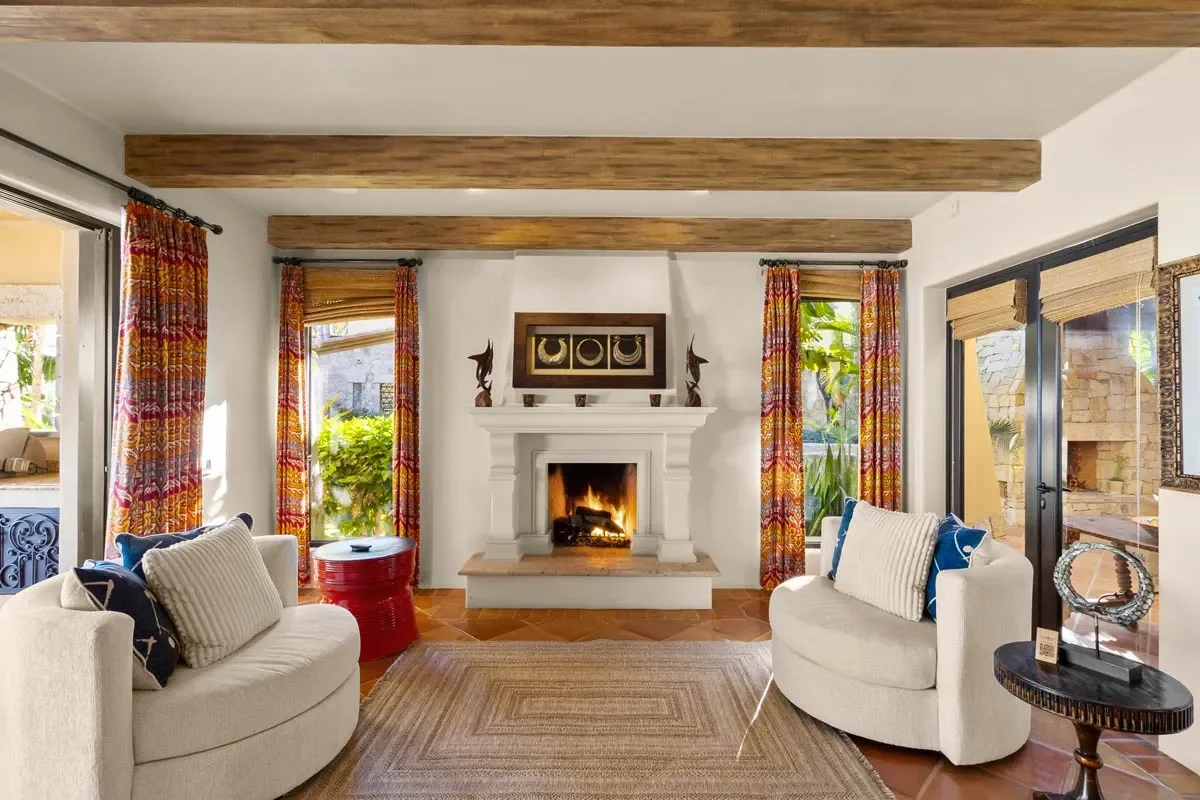 Living room with white armchairs, a fireplace, colorful curtains, and large windows with views of greenery.