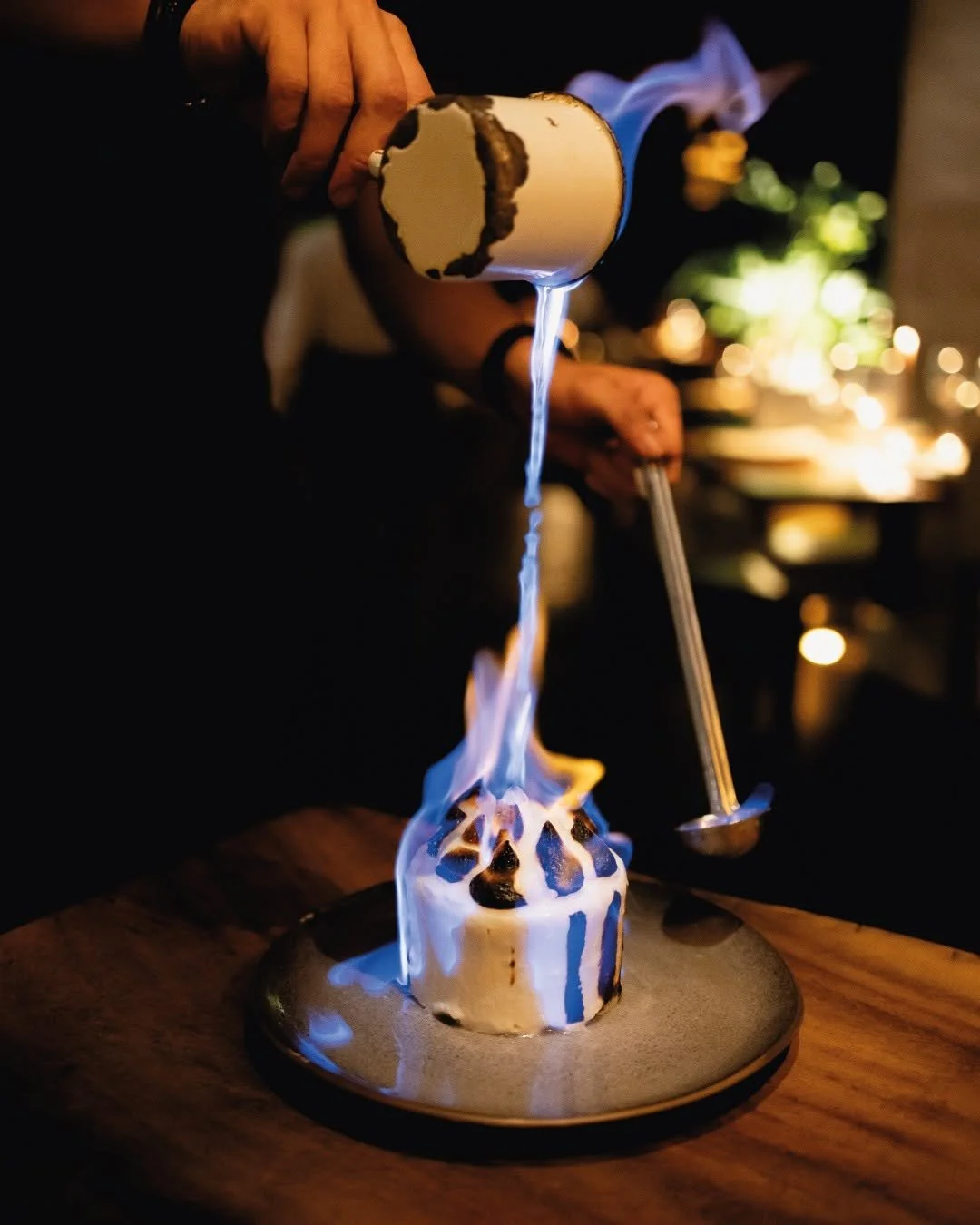 Person pouring a flaming drink into a dessert topped with chocolate and flames, on a dark background with warm bokeh lights.