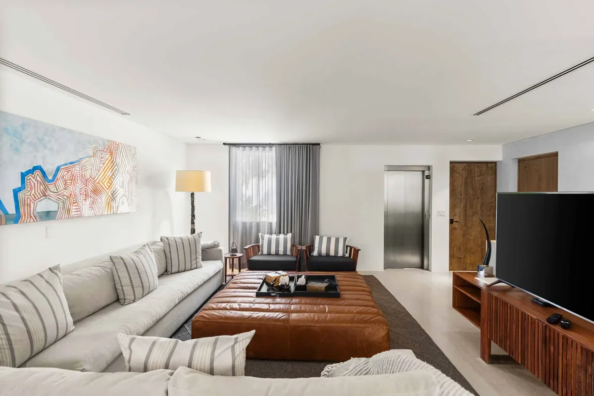 Modern living room with a large beige sectional sofa, striped pillows, a brown leather ottoman with a tray, two black armchairs with striped pillows near a window, a tall floor lamp, a large flat-screen TV on a wooden console, abstract wall art, and 