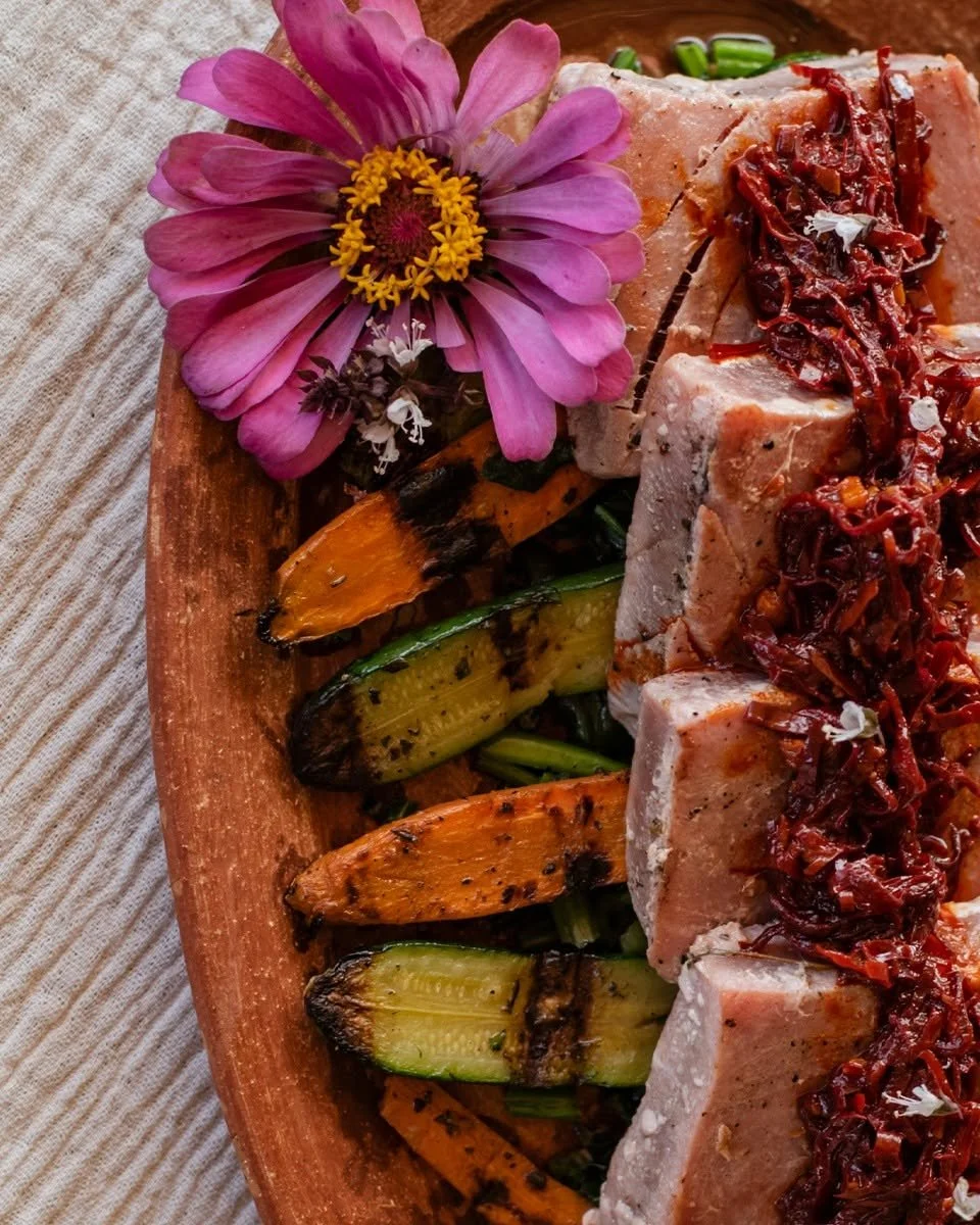 A wooden platter with grilled vegetables and sliced meat topped with caramelized onions, garnished with a pink flower and small flowers.