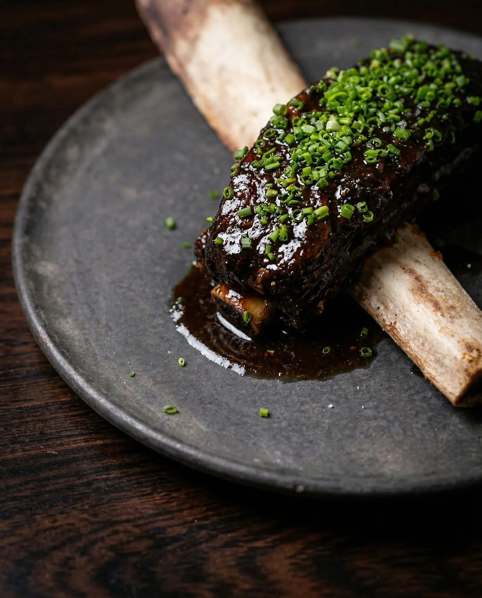 Close-up of a grilled piece of meat topped with chopped green onions and a dark sauce, served on a dark plate with a wooden surface in the background.