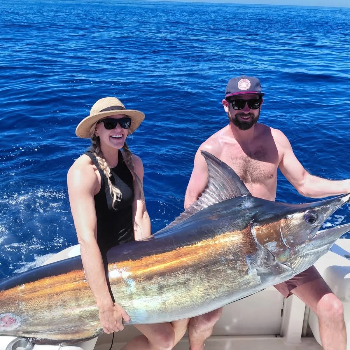 A woman and a man on a boat holding a large fish, with the ocean in the background. The woman is wearing a straw hat and sunglasses, and the man is shirtless with a cap and sunglasses. The fish appears to be a marlin or sailfish.
