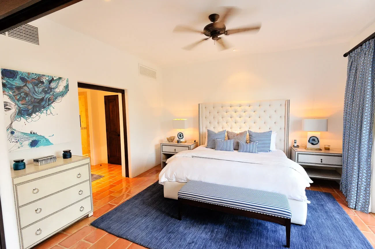 A bedroom with a white upholstered bed with striped pillows, a blue rug, matching nightstands with lamps, a ceiling fan, and a large window with patterned curtains.