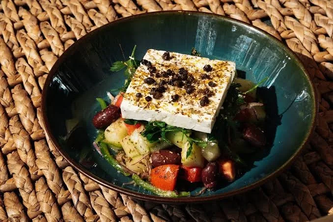 A salad with grilled vegetables and a large piece of cheese topped with black pepper in a green bowl on a woven placemat.