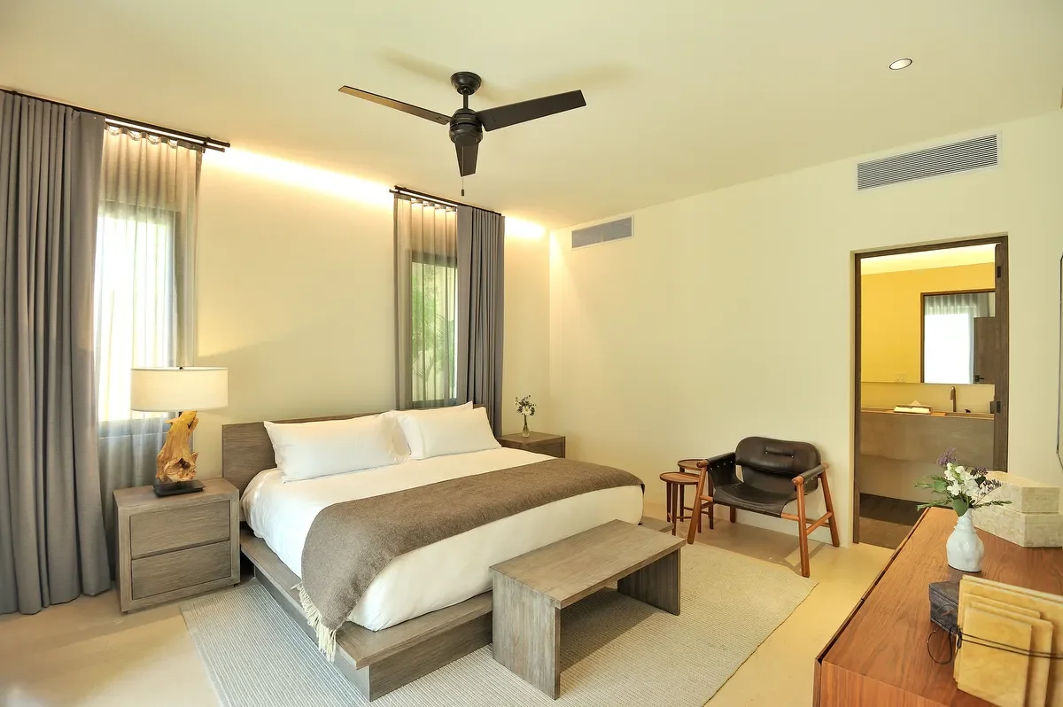 A modern hotel room with a large bed, bedside tables, a ceiling fan, and a small sitting area with two chairs. There are windows with curtains, and a view into a bathroom through an open door.