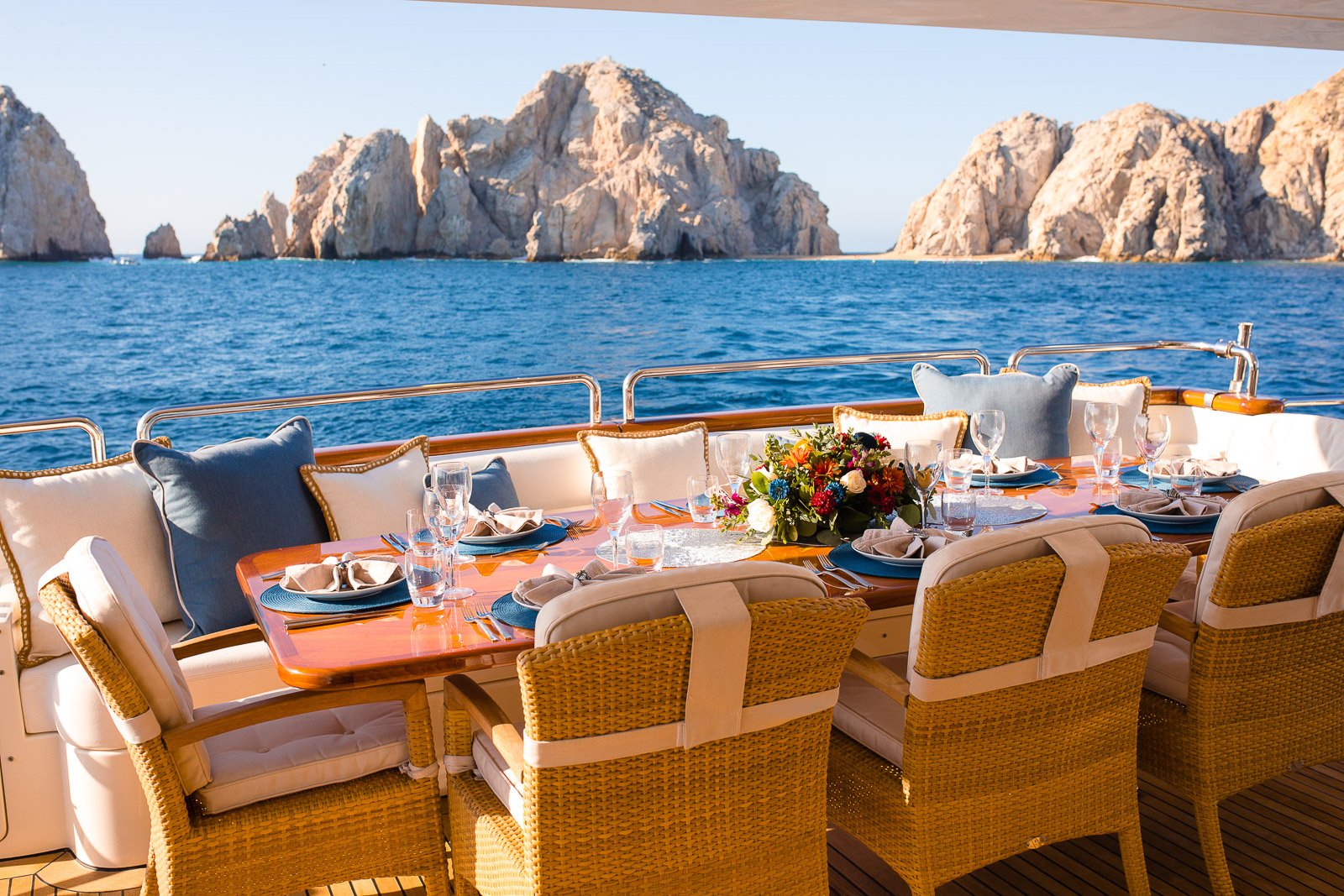 A dining table set with plates, glasses, napkins, and a floral centerpiece on a boat deck with a view of rocky cliffs and the ocean in the background.