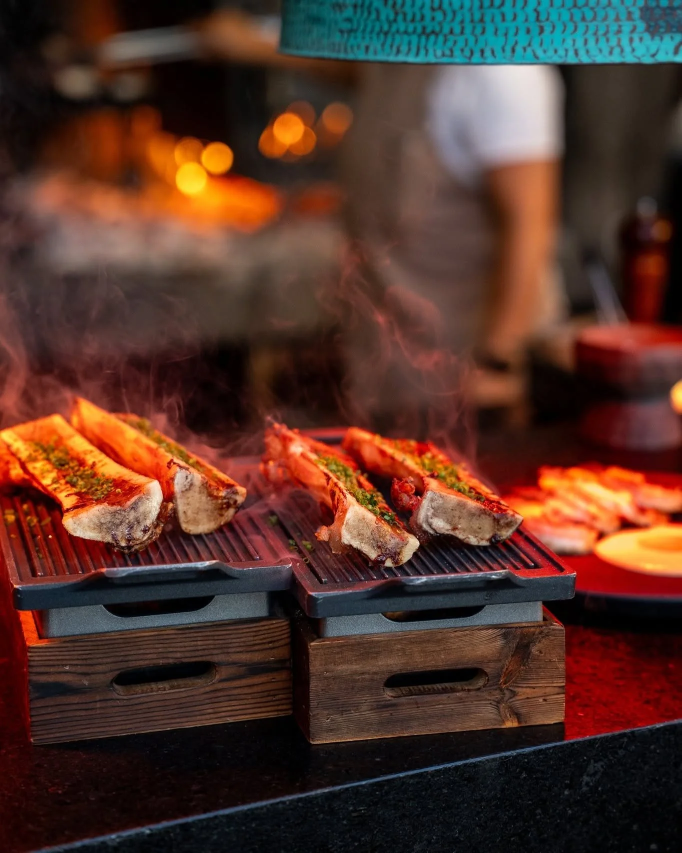 Slices of grilled meat, possibly ribs, on a grill with smoke rising, in a dimly lit setting with warm, yellow lighting.
