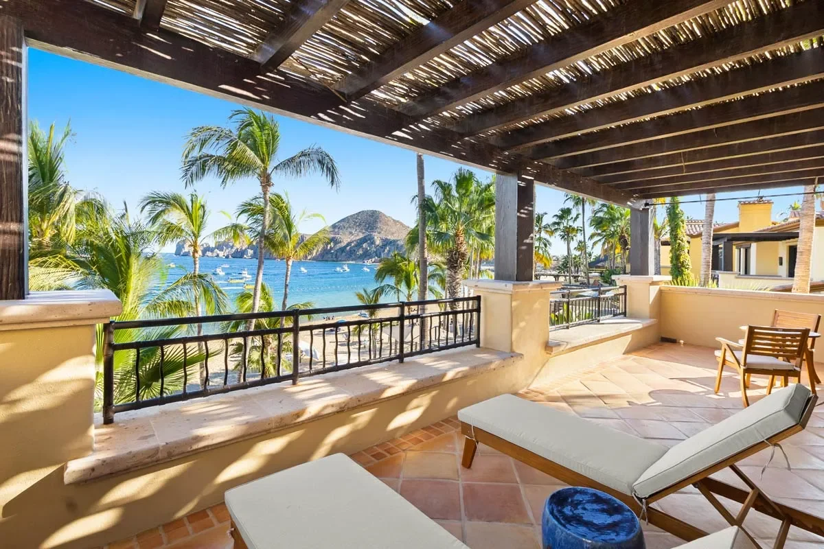 A tropical balcony overlooking a beach and ocean with palm trees, boats, and a mountain in the background, shaded by a wooden pergola.
