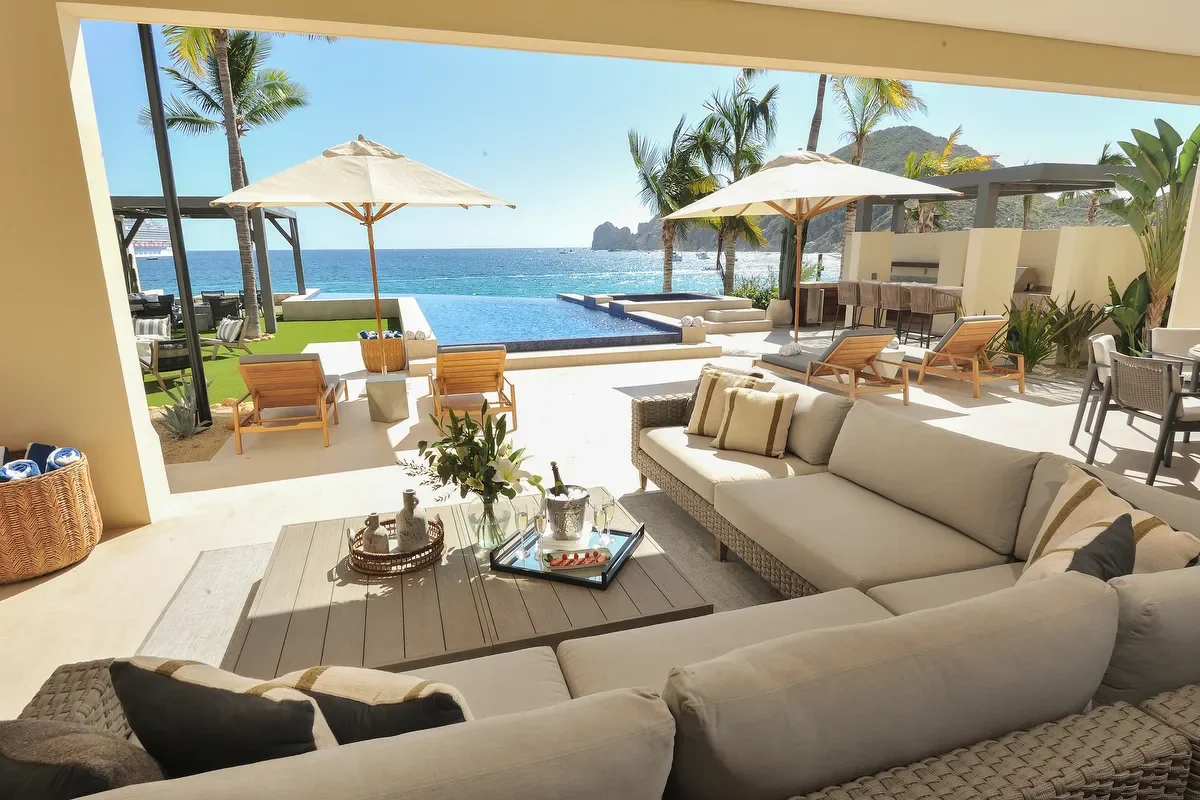 Luxury outdoor patio and pool area with ocean view, sun umbrellas, lounge chairs, palm trees, and seating arrangements under a covered patio.