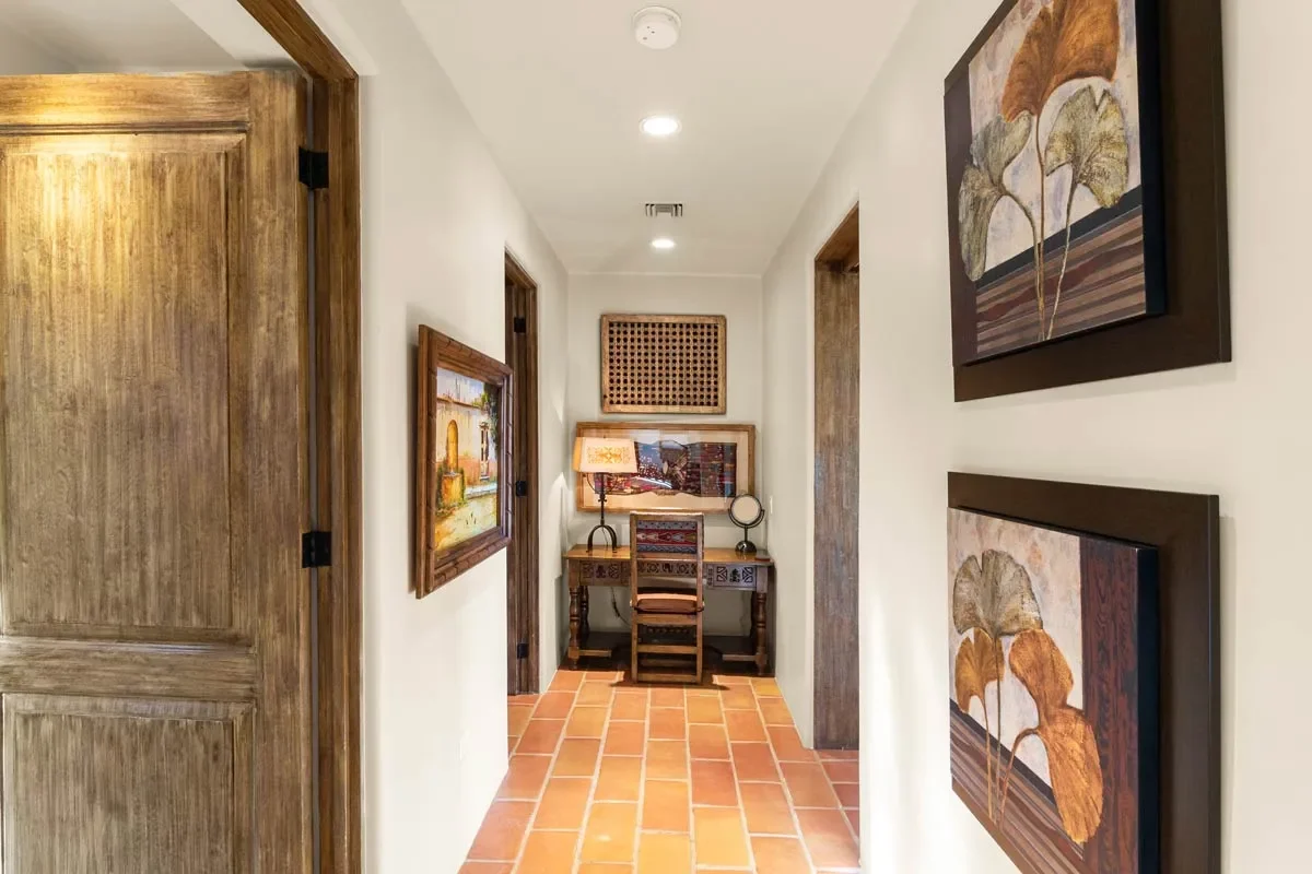A hallway leading to a small desk with a lamp on it, framed artwork on the wall, and terracotta tile flooring.