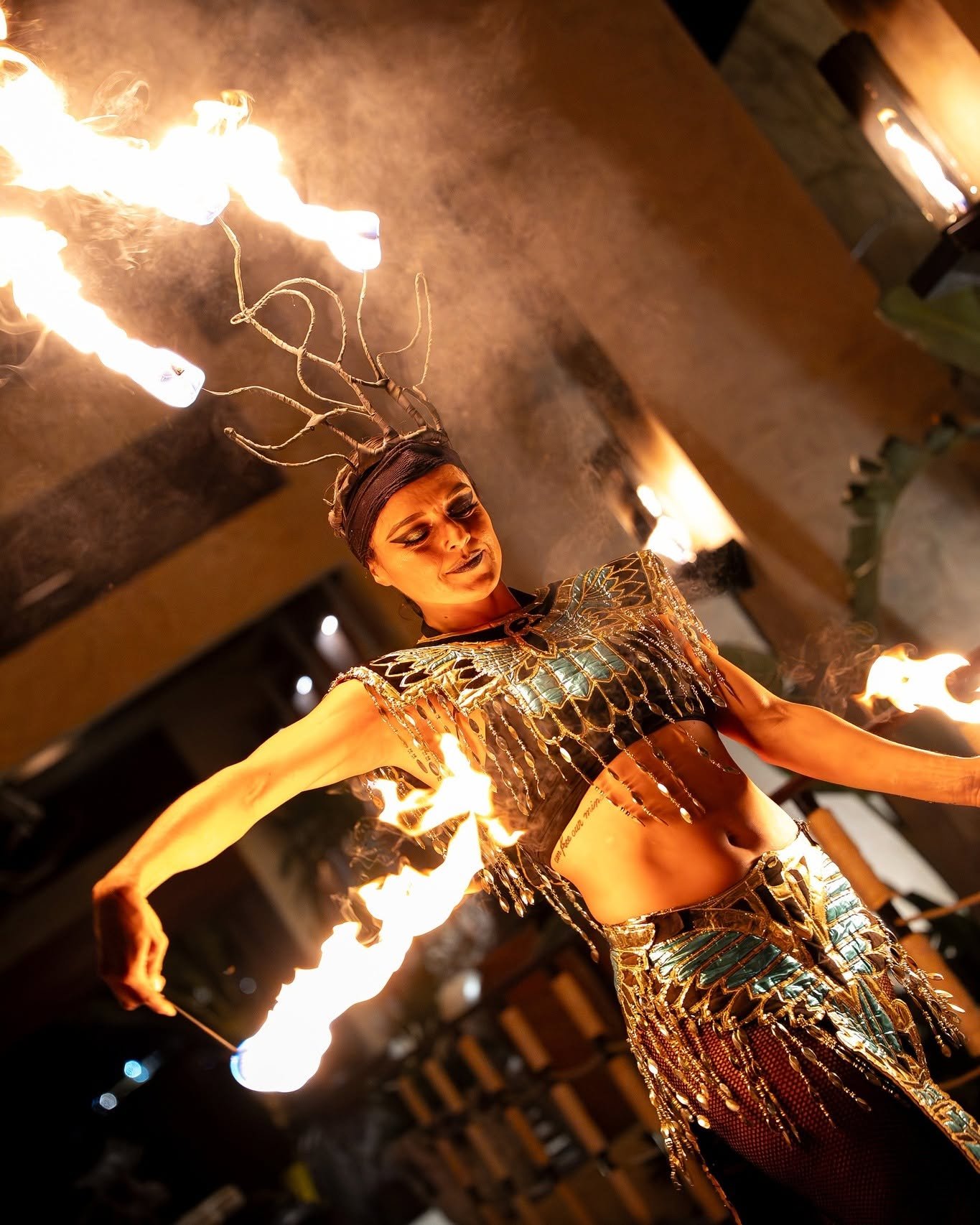 A performer dressed in a detailed, shiny costume with metallic patterns, performing fire spinning with flaming poi or similar tools, in a dimly lit indoor setting.