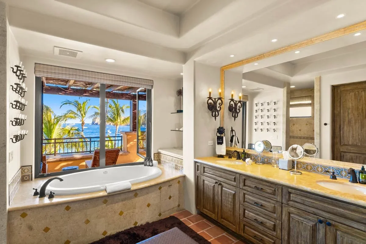 Bathroom with a view of tropical palm trees and water outside the window, featuring a bathtub, large mirror, and double sink vanity.