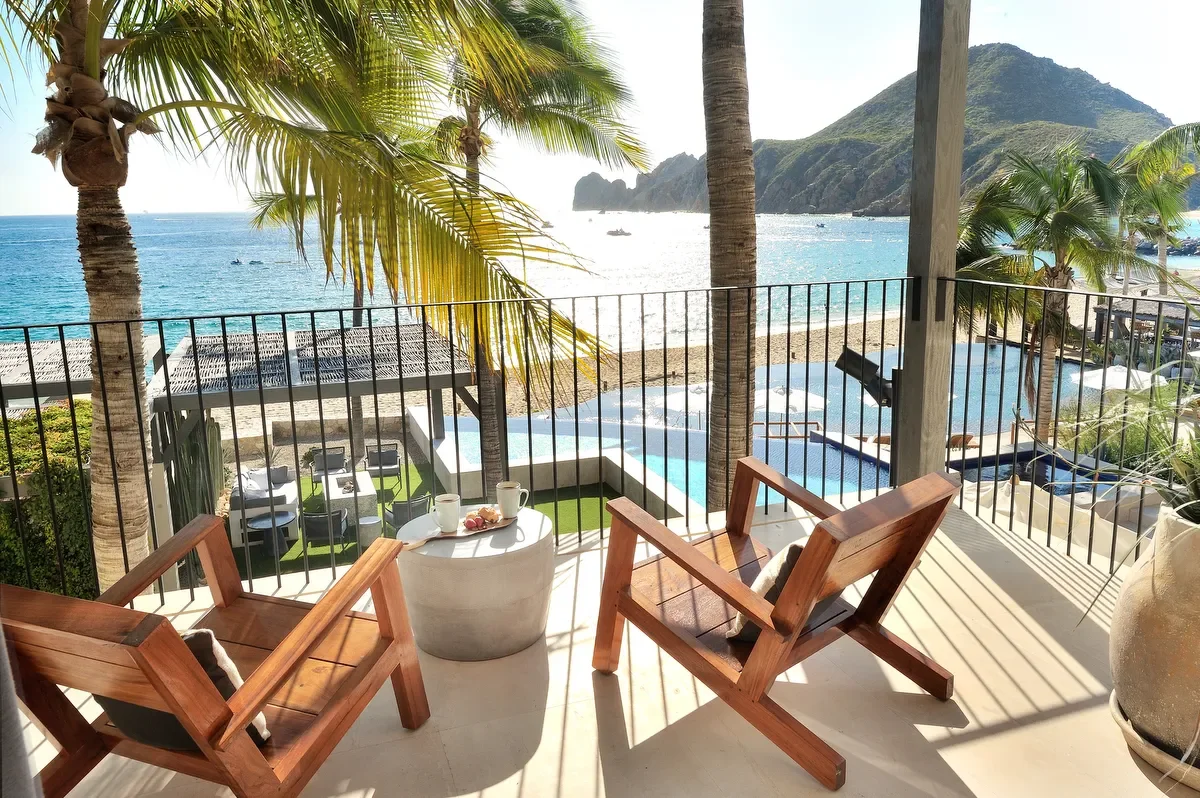 Balcony overlooking a beach with palm trees, outdoor seating, and a swimming pool, with mountains in the background.