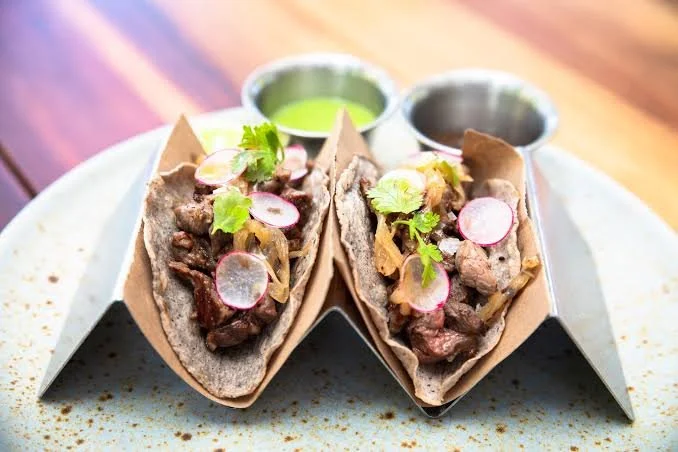 Two tacos filled with cooked meat, topped with radish slices, onions, and cilantro, served with green and dark sauces.