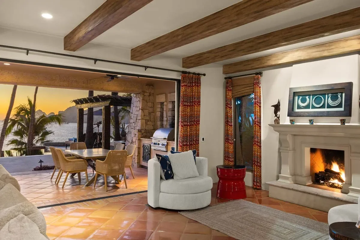 Living room with fireplace, white armchair with pillows, red side table, colorful curtains, open patio with dining table, chairs, barbecue grill, ocean view at sunset, palm trees, stone wall, wood beam ceiling.