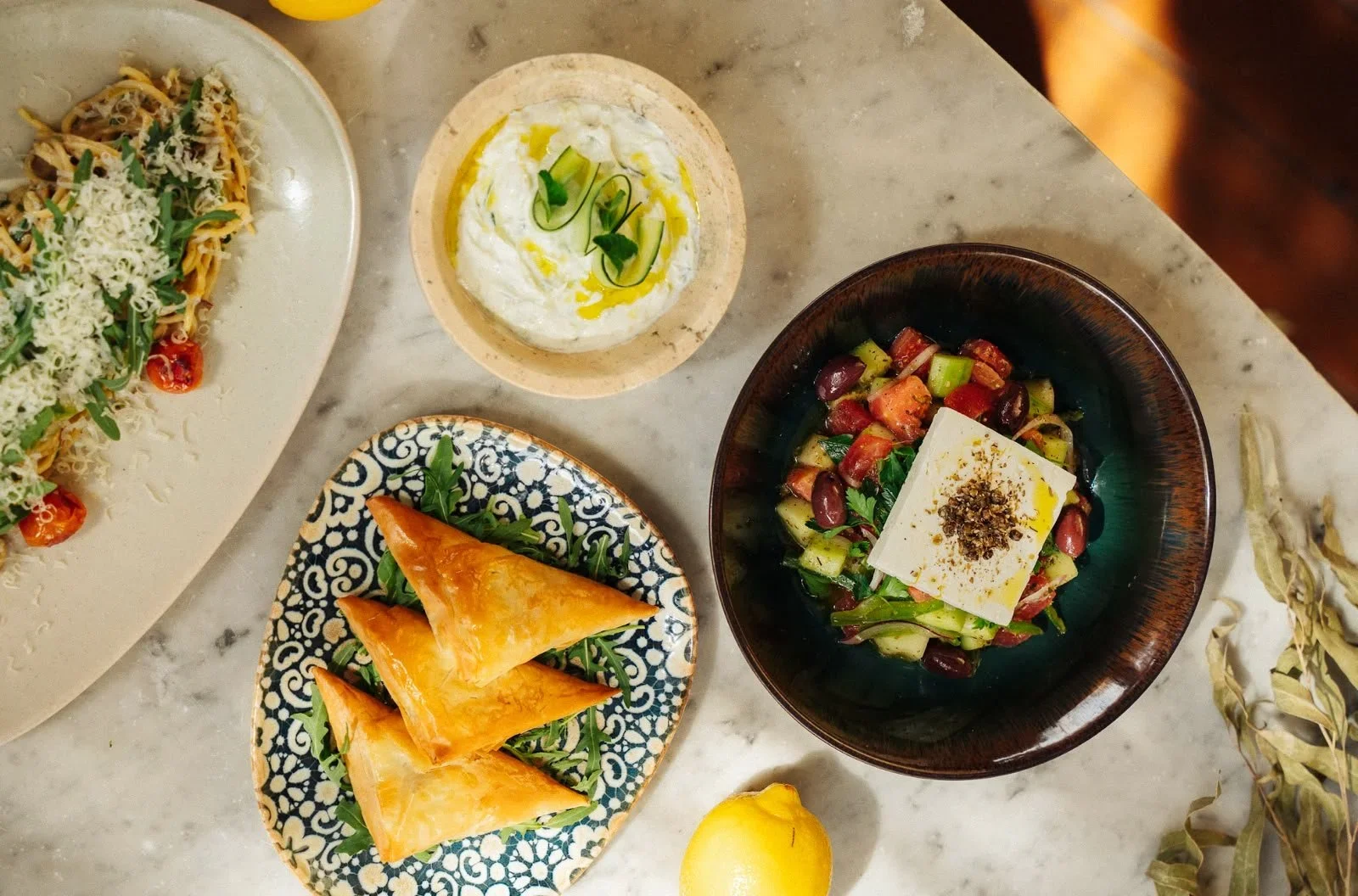 Assorted Mediterranean dishes on a marble table, including a salad with cubed vegetables and cheese, a plate of samosas, a bowl of yogurt with cucumber and herbs, and a pasta dish with cheese and vegetables.