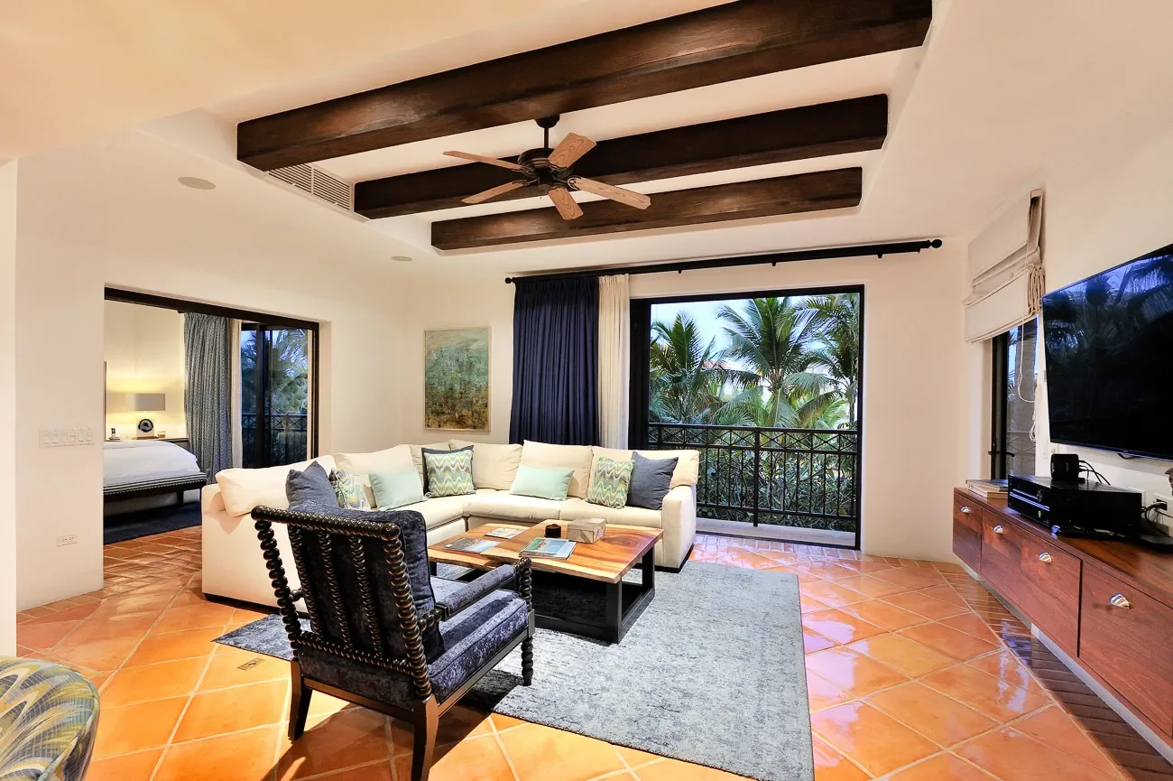 Living room with a white sectional sofa, dark armchair, wooden coffee table, large sliding glass door with view of palm trees, flat-screen TV, and a bedroom visible through a doorway, all on terracotta tile flooring.