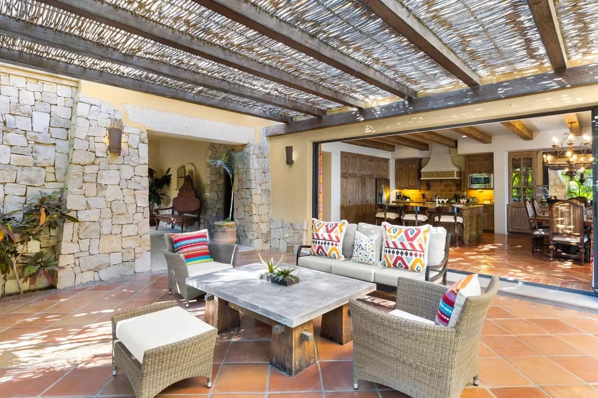 Outdoor covered patio with stone walls, terracotta floor tiles, and a wooden pergola roof. The space includes outdoor seating with colorful pillows around a rectangular coffee table. The background shows an open kitchen and dining area with wooden ca