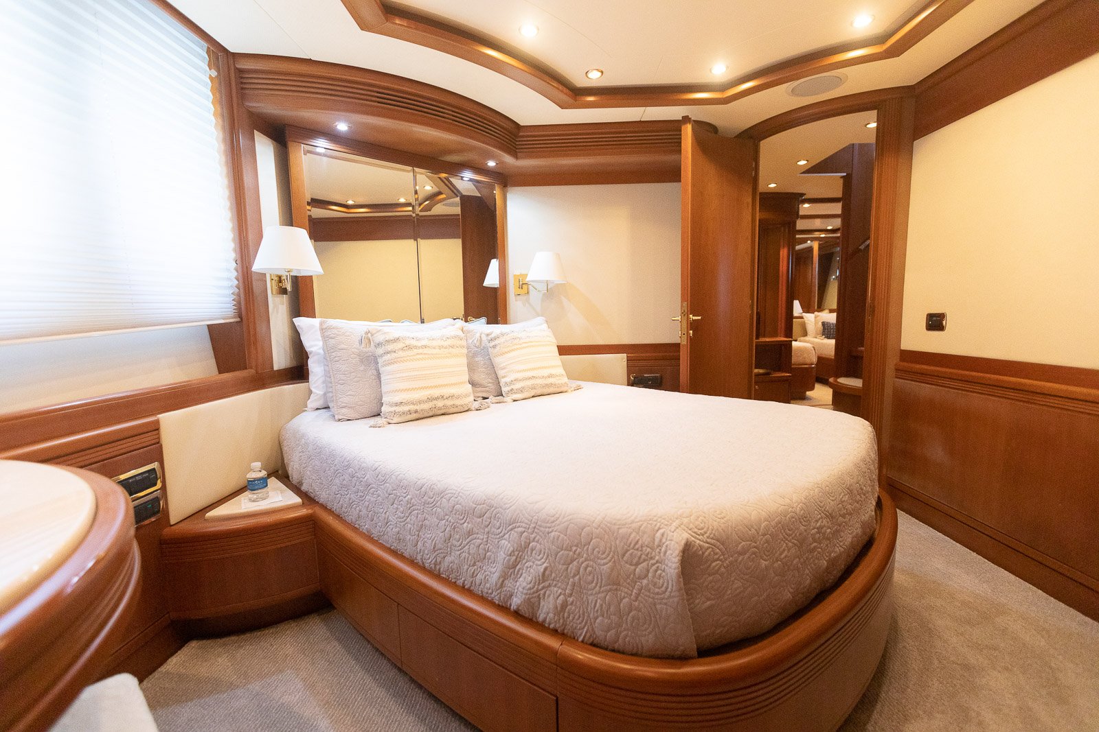 Luxurious hotel bedroom with a large bed, multiple pillows, wooden trim, and a mirror. There's a window with blinds on the left and a door on the right leading to a sitting area.