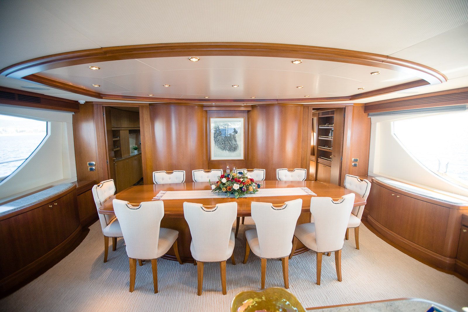 Elegant dining room with a large wooden table, nine upholstered chairs, floral centerpiece, framed artwork, wood-paneled walls, built-in cabinets, and large windows.