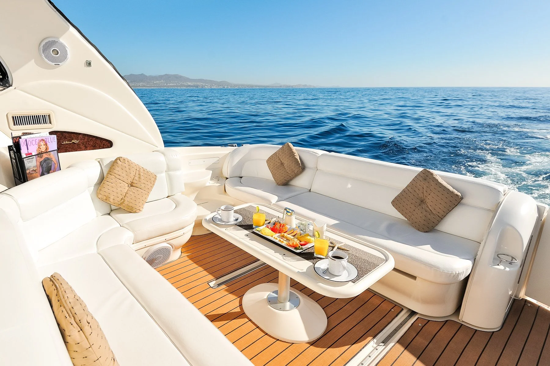 Luxury yacht deck with white cushioned seating, pillows, a coffee table with breakfast items, drinks, and a view of the open ocean and distant mountains.
