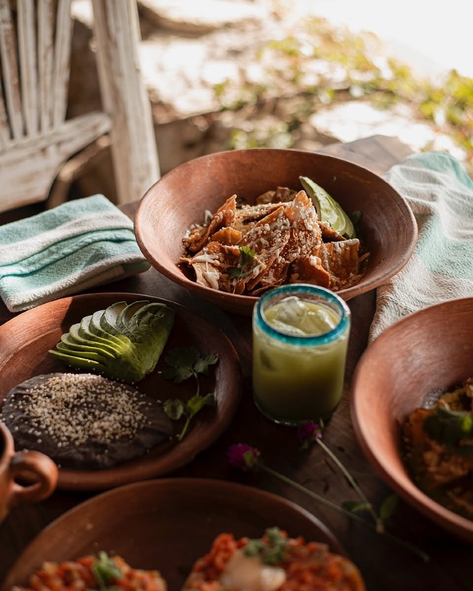 A traditional Mexican meal on a rustic wooden table featuring bowls of shredded meat, sliced avocado, and a green beverage, with a colorful outdoor background.