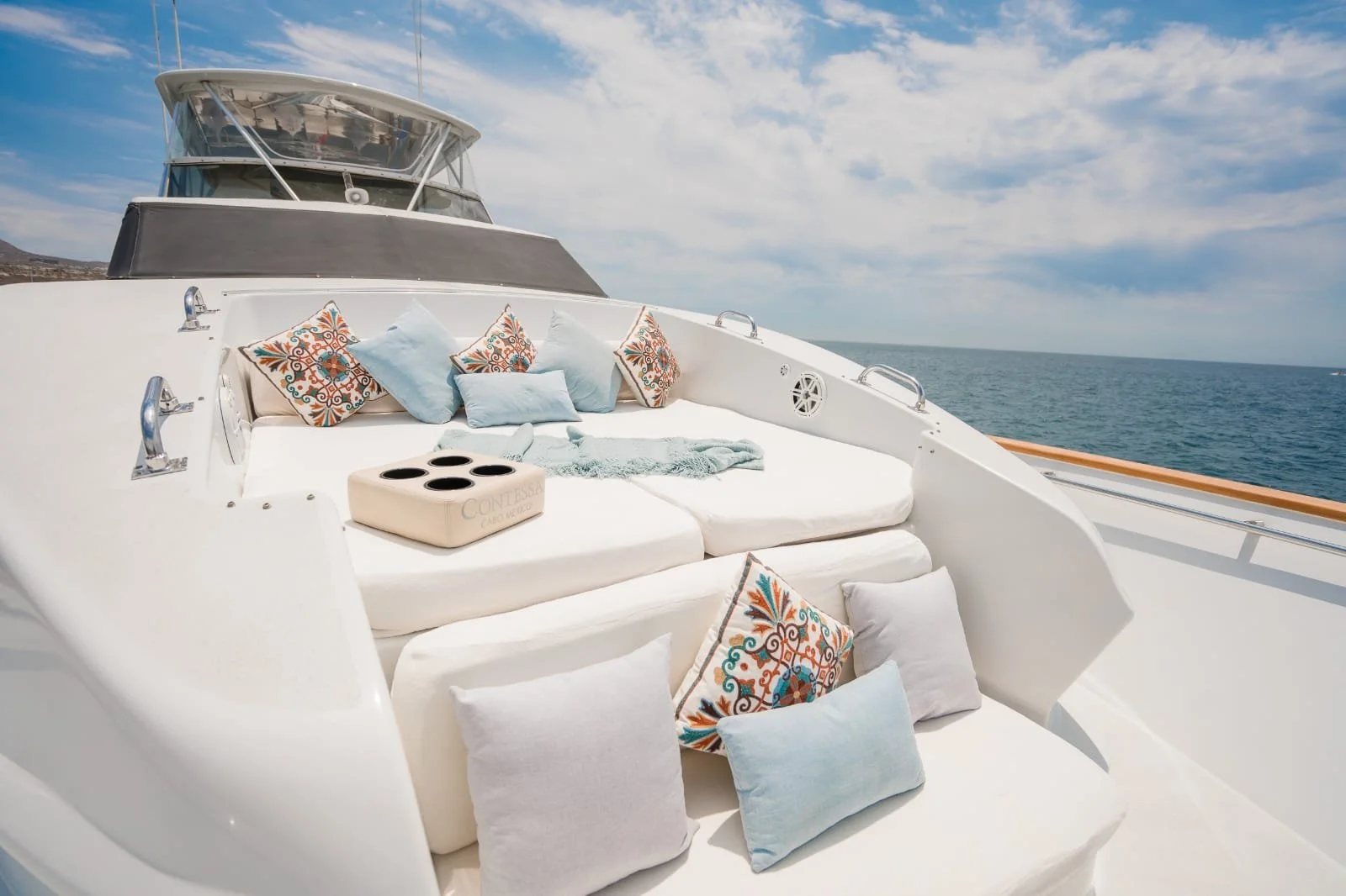 Deck of a yacht with white cushions, colorful patterned and light blue pillows, a blanket, and a beige cornhole game box with black holes, overlooking the ocean under a partly cloudy sky.
