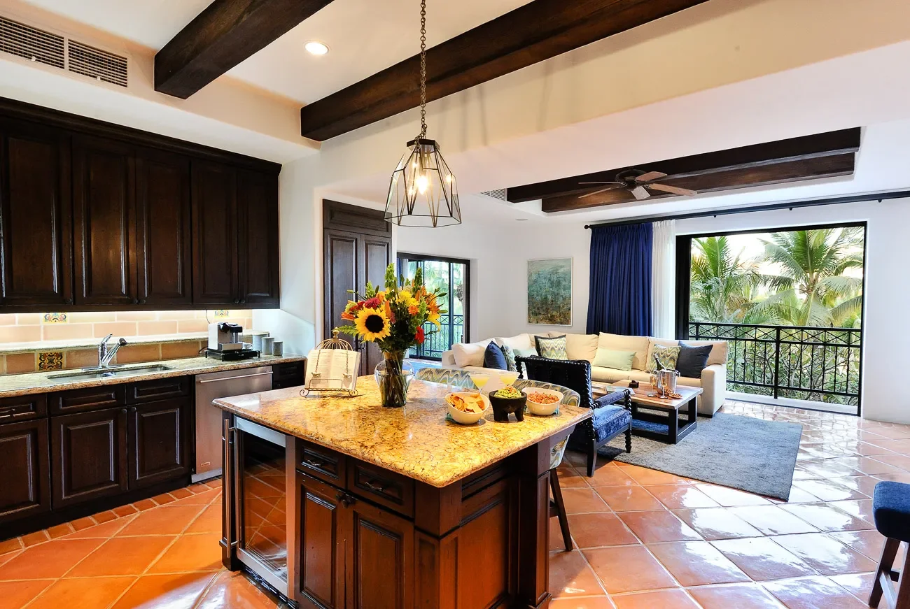 Open-concept kitchen and living room with dark wood cabinets, granite countertop island, a bouquet of colorful flowers, and a seating area with white sofa and blue curtains, overlooking green palm trees outside.