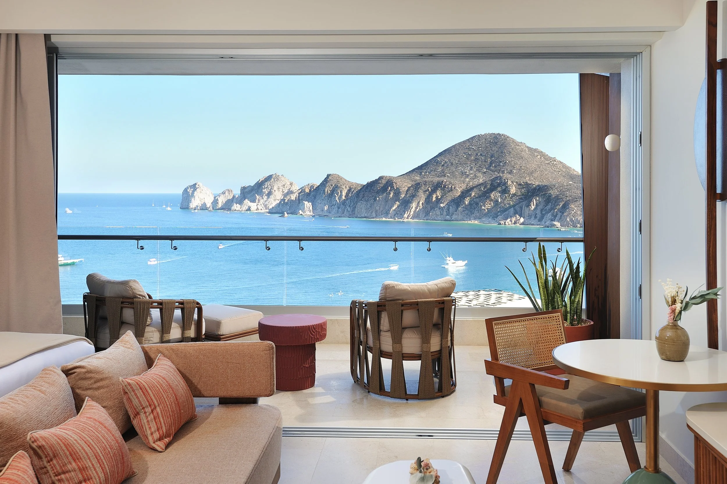 A living room with a large window showing a view of rocky cliffs and the ocean with boats. The room has beige sofas with red and beige pillows, a round white table with a small plant, and wooden chairs with woven backrests. There is a potted plant near the window and a pink cylindrical side table.
