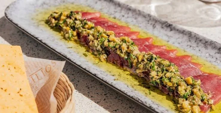 A rectangular plate of sliced raw tuna covered with chopped pistachios and herbs, drizzled with olive oil, on a speckled countertop.