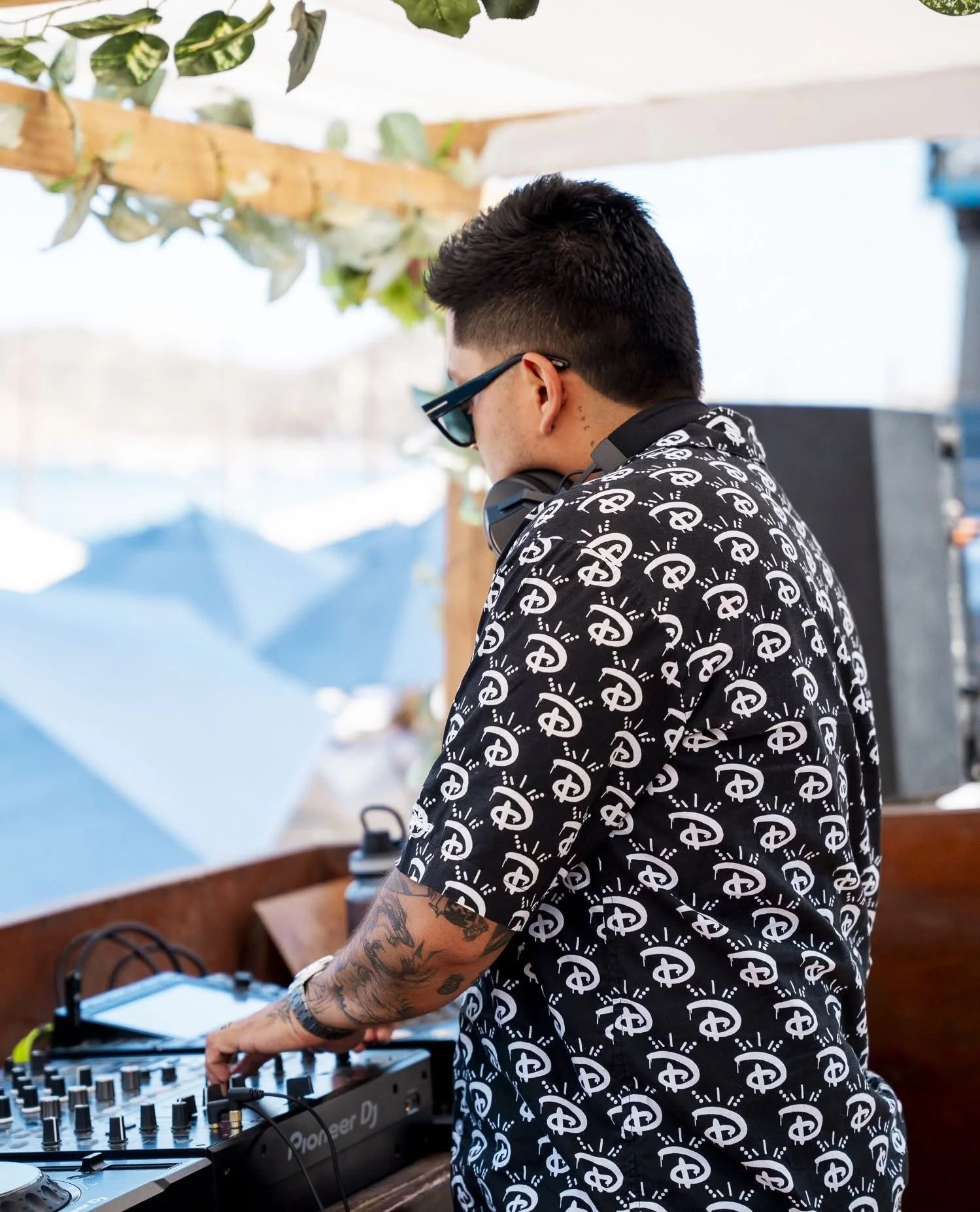 Side profile of a man with tattoos on arm, wearing sunglasses and a shirt with a dollar sign pattern, standing at a DJ console outdoors.