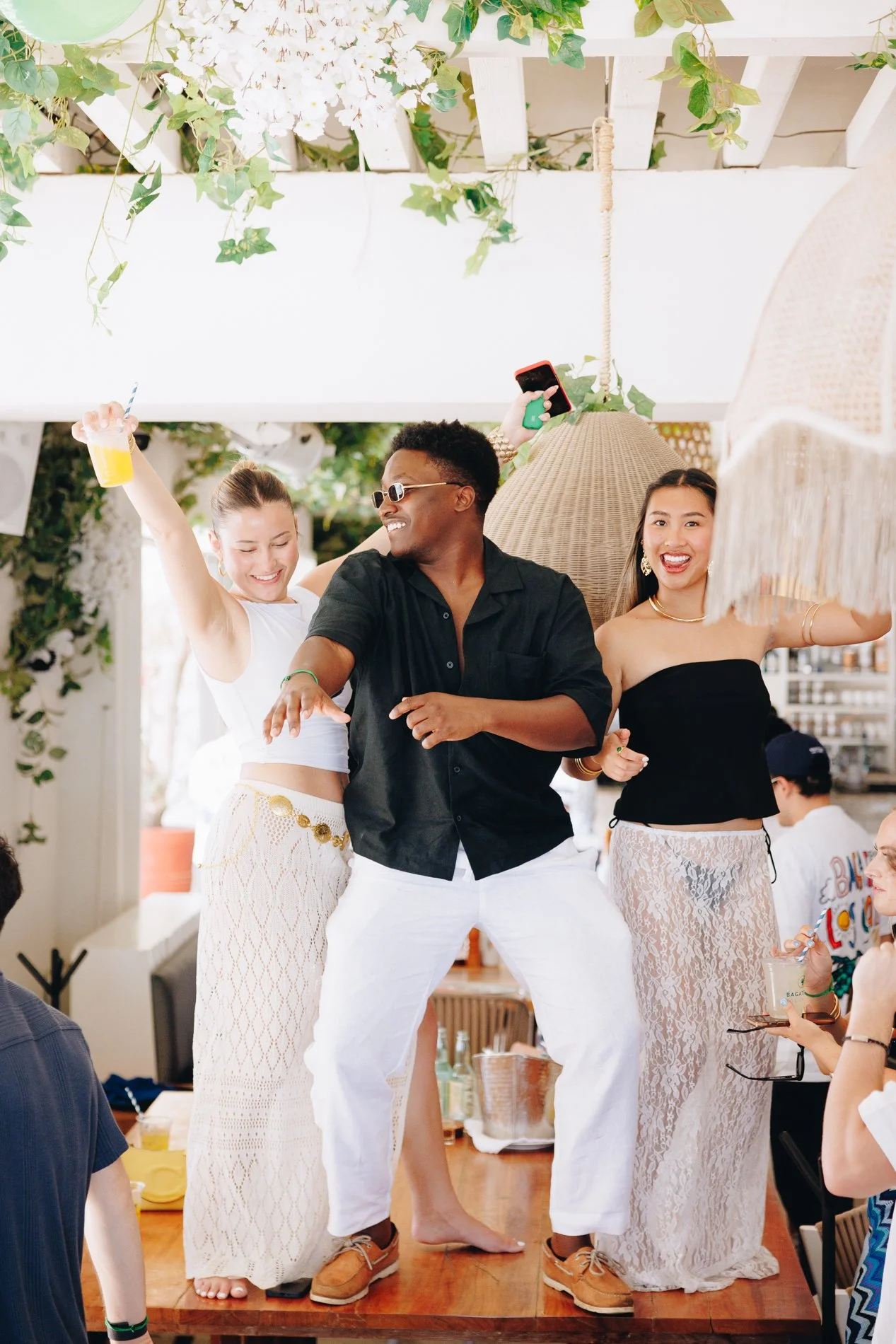 Three people dancing and celebrating at a party. They are smiling, dressed in summer clothes, and appear to be having fun in a decorated indoor space with hanging greenery.