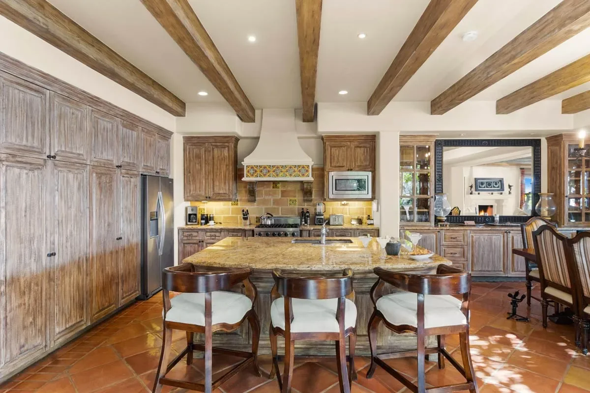 Kitchen with wooden cabinets, a center island with a granite countertop, four barstools, stainless steel appliances, and a visible adjoining living room with a fireplace.