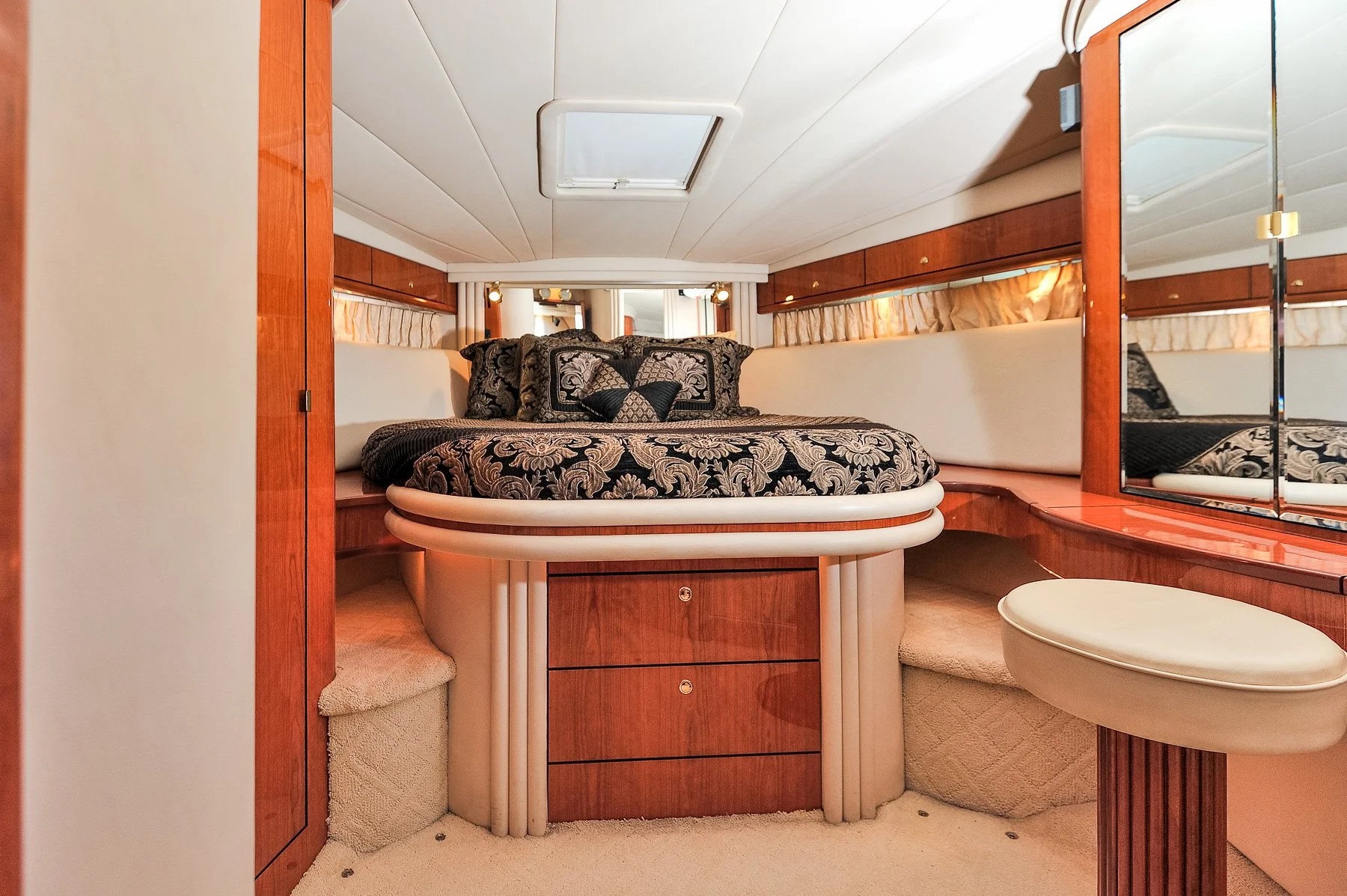 A bedroom inside a boat with a circular bed decorated with black and gold patterned bedding and matching pillows, wood paneling, a skylight, and a large mirror.