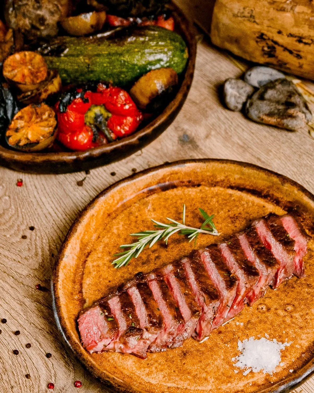 Sliced grilled steak garnished with rosemary on a rustic plate, served with a side of roasted vegetables including zucchini, tomatoes, and eggplant on a wooden surface.