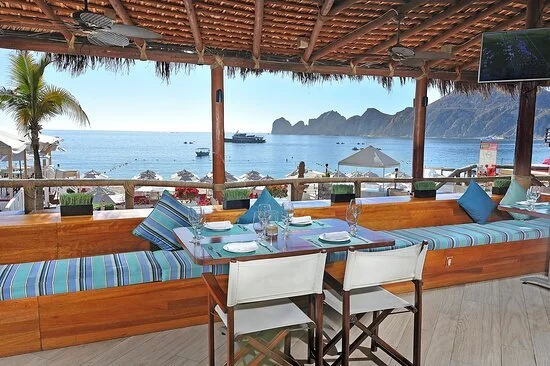 Seaside restaurant with a long wooden table set with menus, plates, glasses, and napkins, overlooking the ocean and rocky cliffs, with a thatched roof and a palm tree on the left.