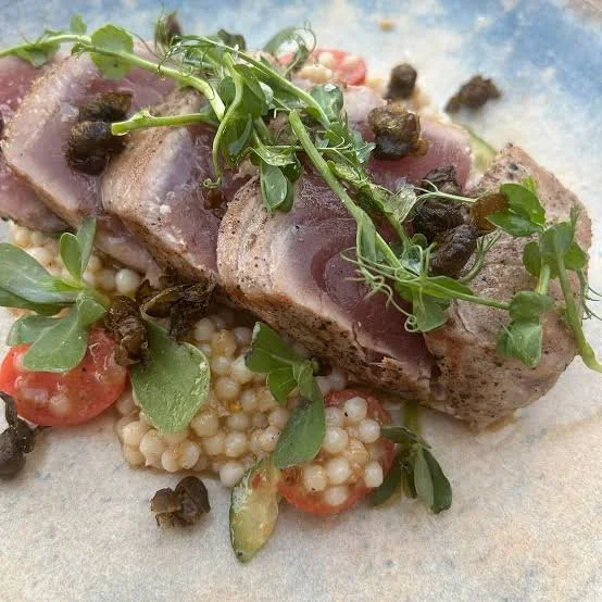 Seared tuna steak topped with microgreens, black caviar, and fresh vegetables, served with a bed of couscous and sliced tomatoes.