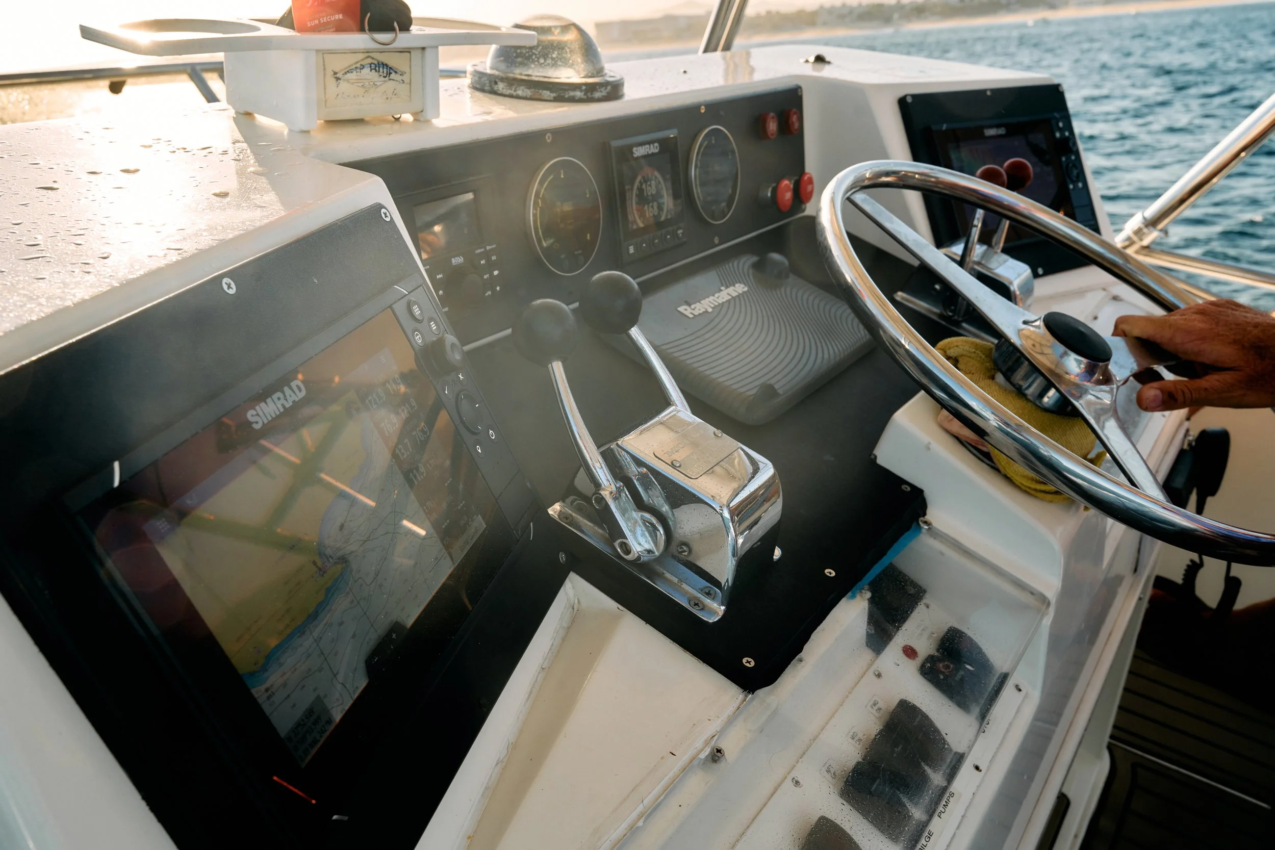 Close-up view of a boat's steering console with navigation and engine controls, displaying water and part of the boat's deck in the background.