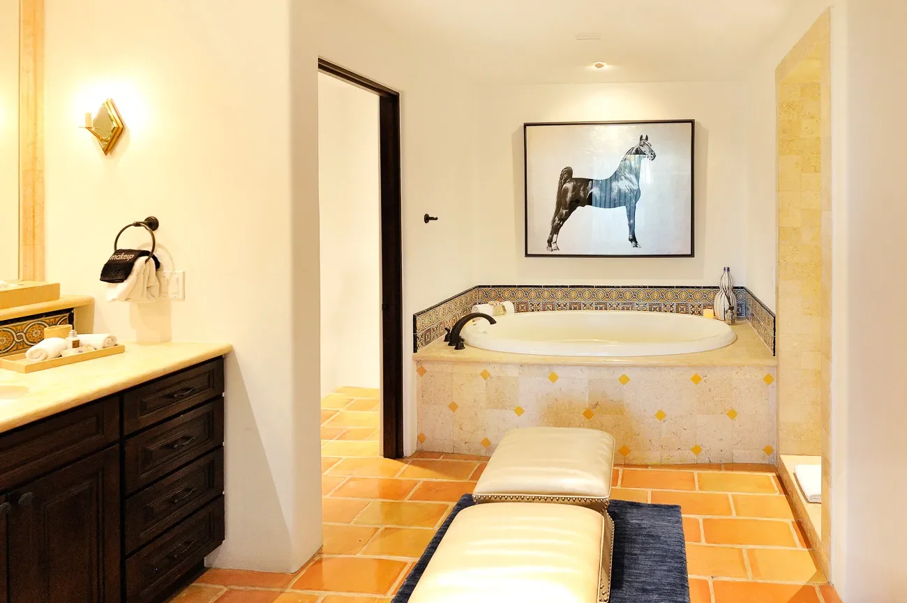 Bathroom with a built-in bathtub decorated with patterned tiles, a lid-covered bench in front, a framed horse artwork on the wall above the tub, a dark wooden cabinet with toiletries, and a small open shower area to the right.