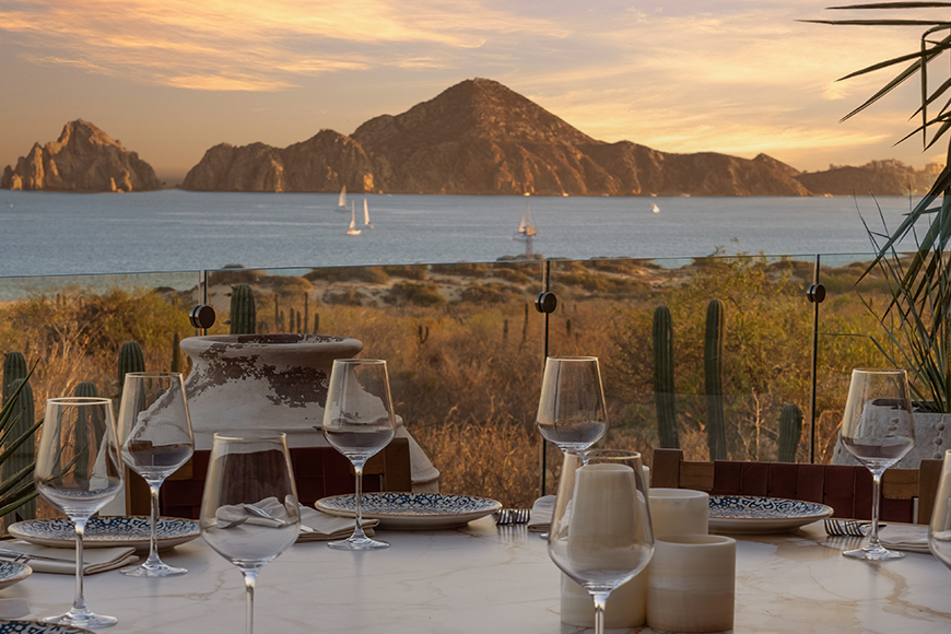 A dining table set with wine glasses, plates, and candles on an outdoor terrace with a view of desert landscape, cacti, and mountains with sailboats on water in the background during sunset.