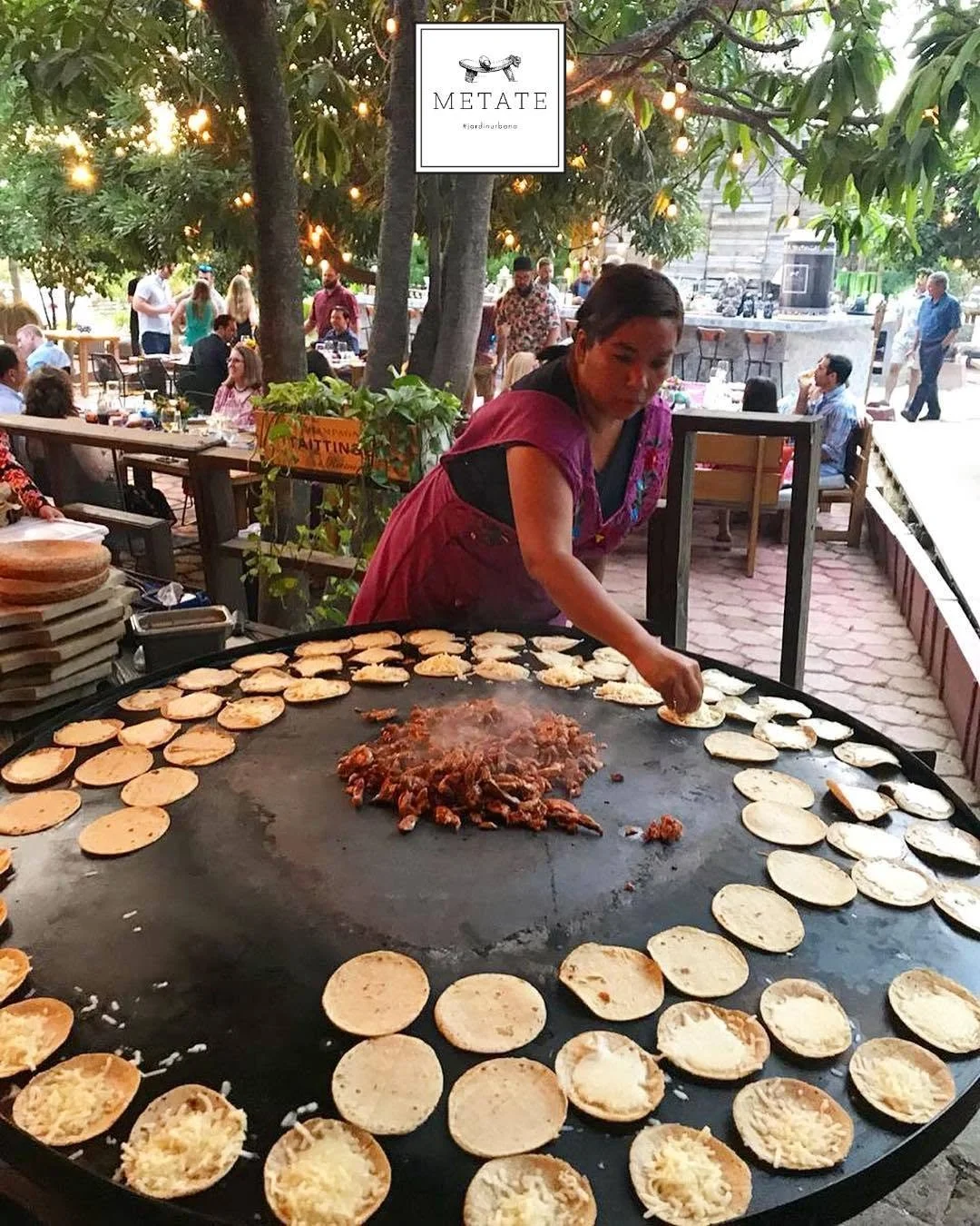 A woman cooking Mexican tacos on a large outdoor griddle with rows of tortillas, surrounded by trees and people dining at tables in a restaurant patio.