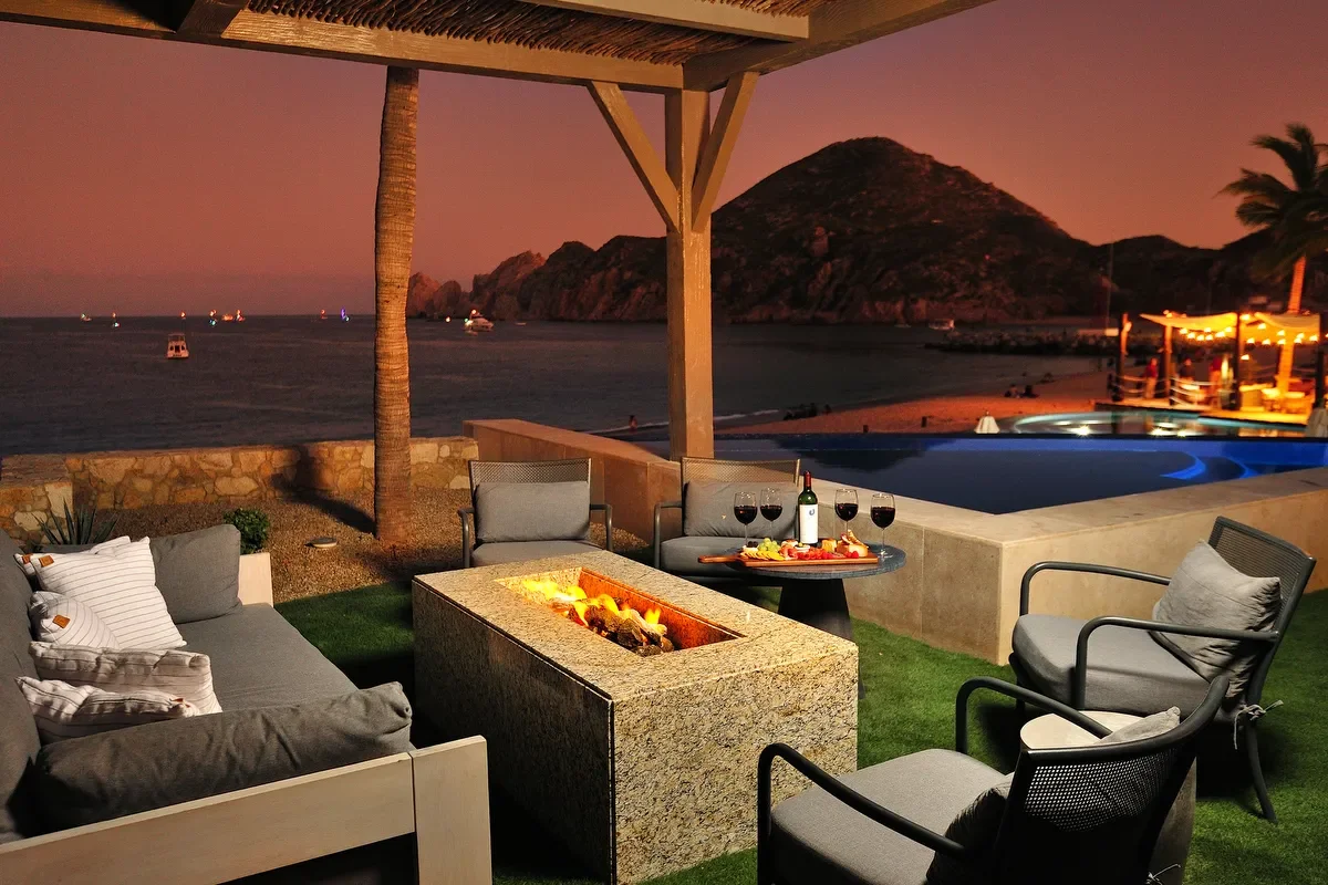 Outdoor seating area with pillows, a fire pit, a small table with wine and snacks, near a pool overlooking the ocean and a mountainous island at sunset.