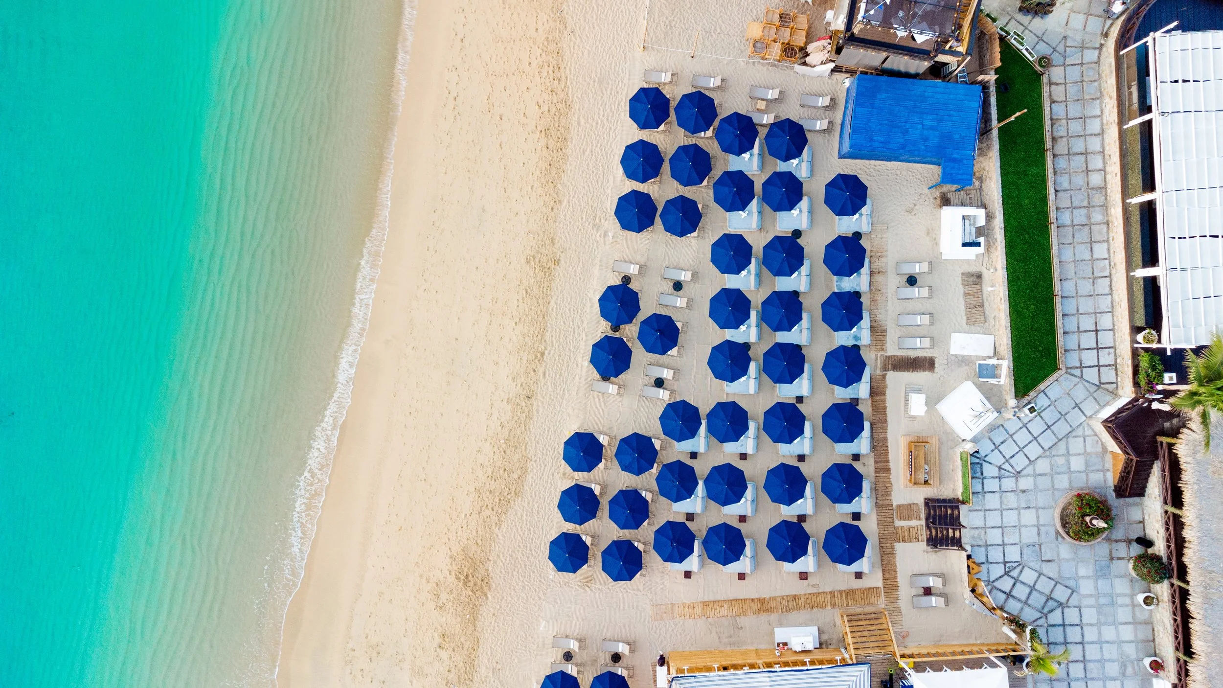 Aerial view of a beach with turquoise water, sandy shoreline, and multiple blue beach umbrellas with lounge chairs.