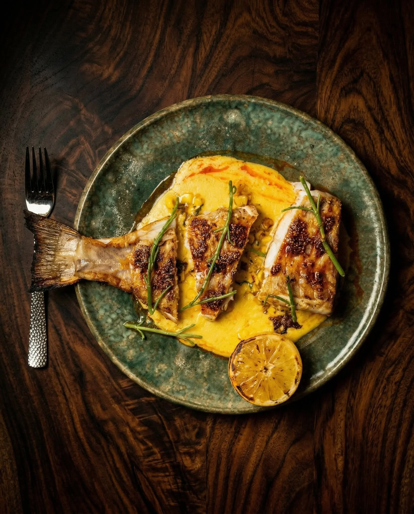 A cooked fish fillet topped with mashed potatoes, garnished with green herbs, served on a rustic ceramic plate. A halved grilled lemon is on the side on a dark wooden table.