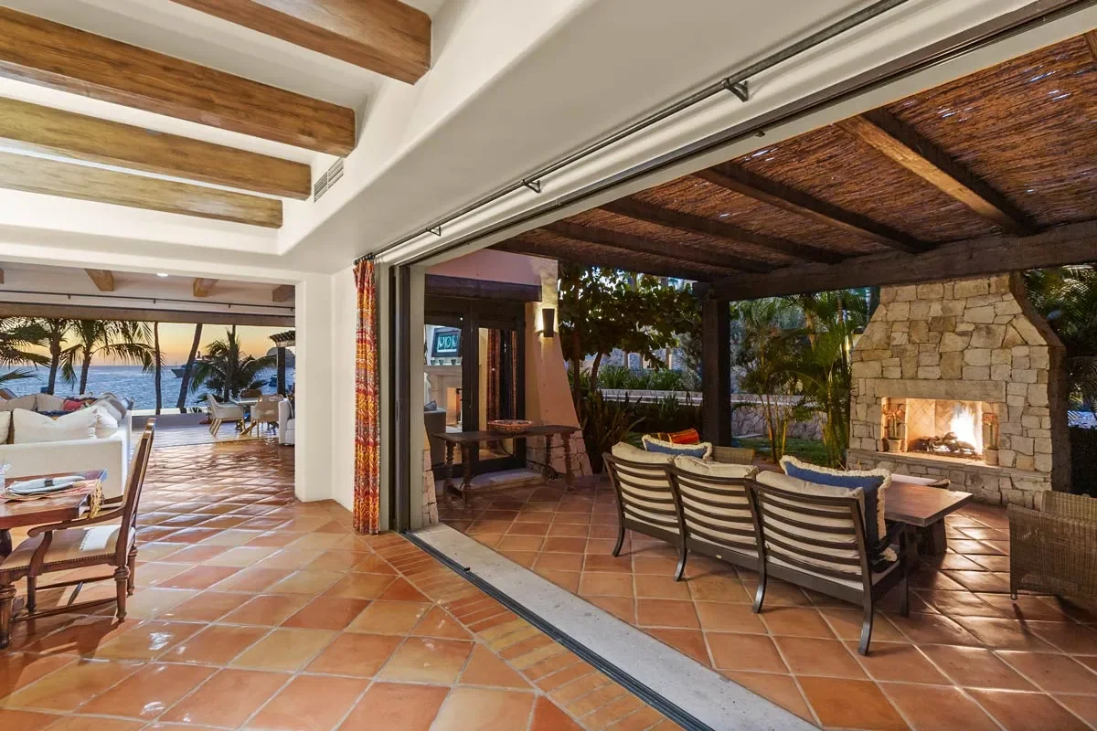 Indoor-outdoor living area with terracotta tile floors, wooden beams, and a stone fireplace, opening to a patio with outdoor furniture and a view of palm trees and the ocean at sunset.