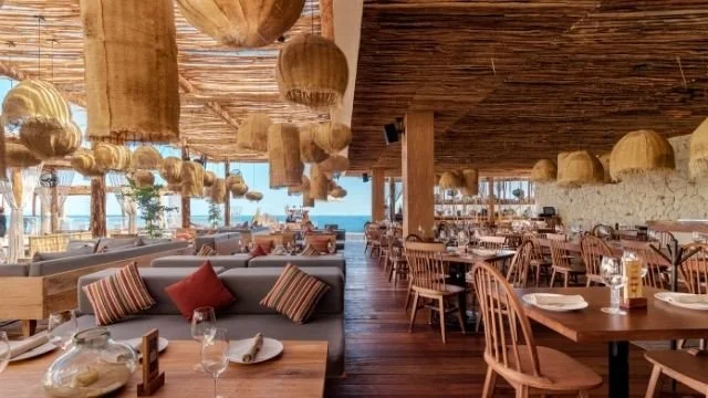 Interior of a beachside restaurant with wooden furnishings, hanging pendant lights, and tables set for dining.