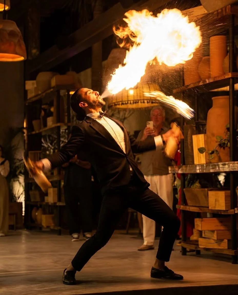 A man in a tuxedo is performing a fire-breathing act indoors, with flames coming out of his mouth.