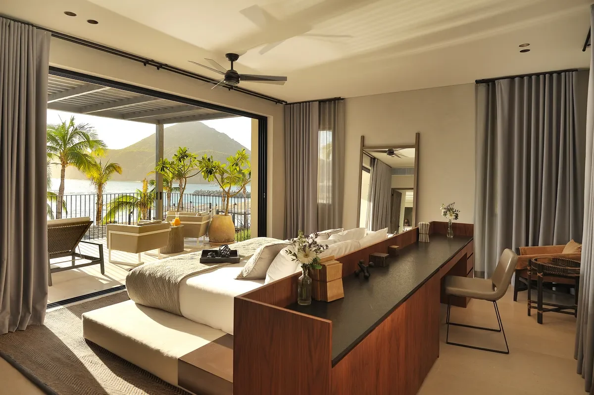 Living room with large open sliding door leading to balcony, ocean view, tropical plants, mountain in the background, white sofa, balcony chairs, and a reflection of the room in a tall mirror.